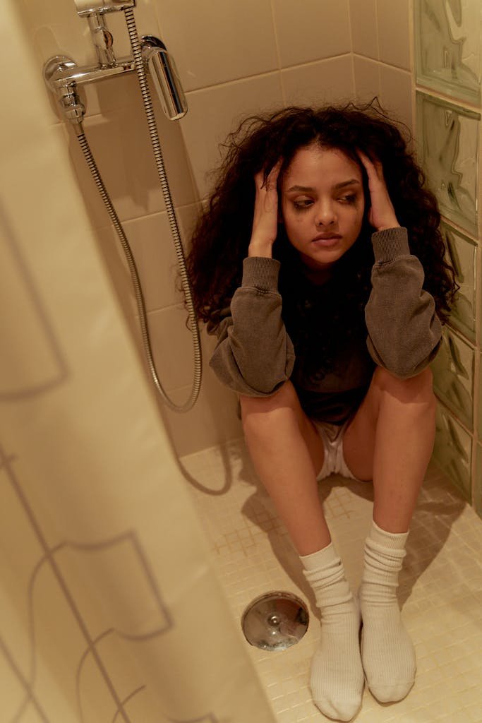 A young woman with curly hair sits thoughtfully in a shower, reflecting on her emotions.