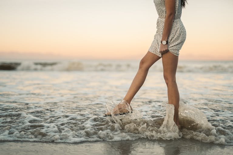 A woman in a dress splashes through the ocean waves at sunset, creating a serene beach moment.